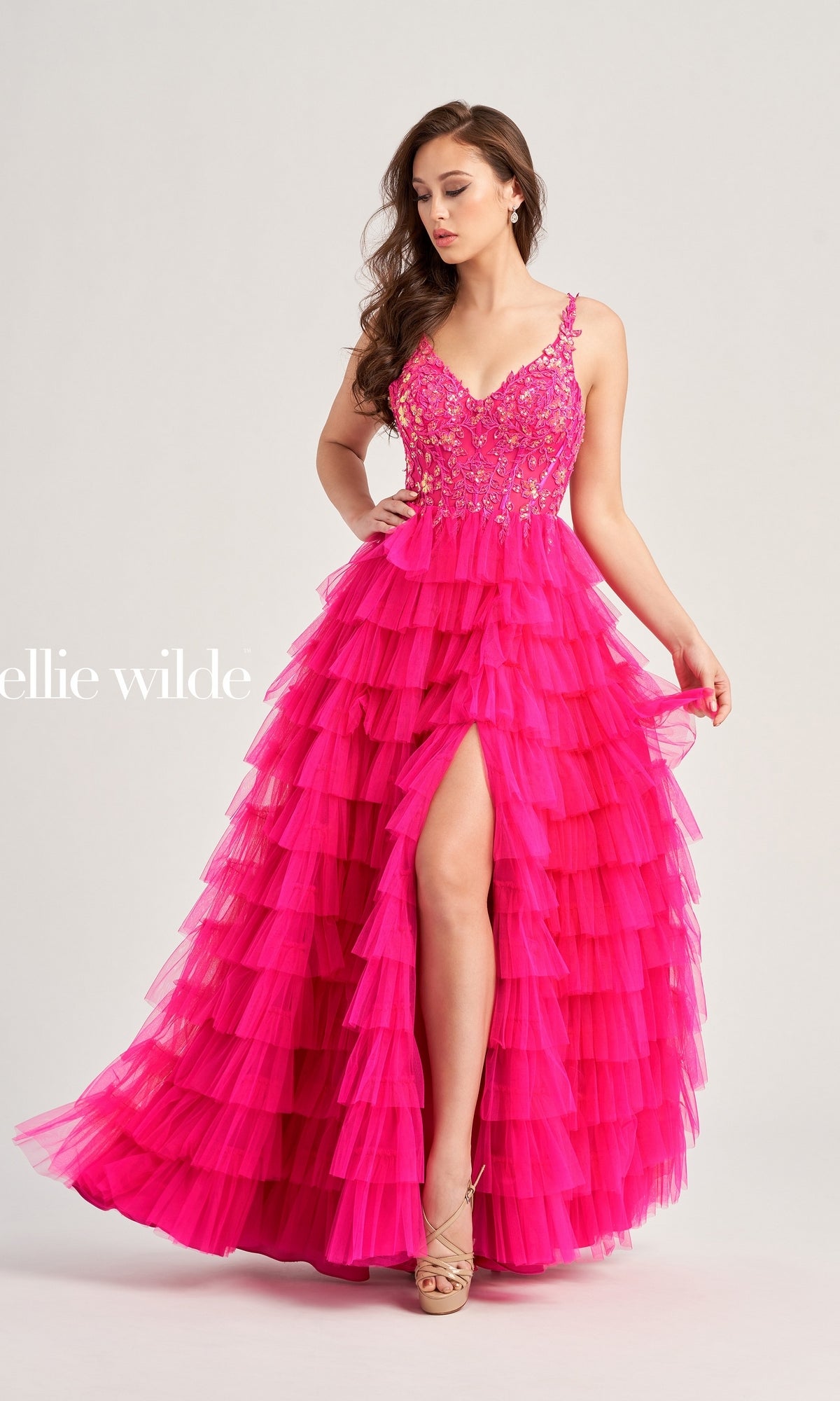 A woman wearing the Tiered Ellie Wilde EW35059 Designer Ball Gown with a sequin corset bodice, ruffled skirt, thigh-high slit, floral details, and nude heels stands against a plain background.